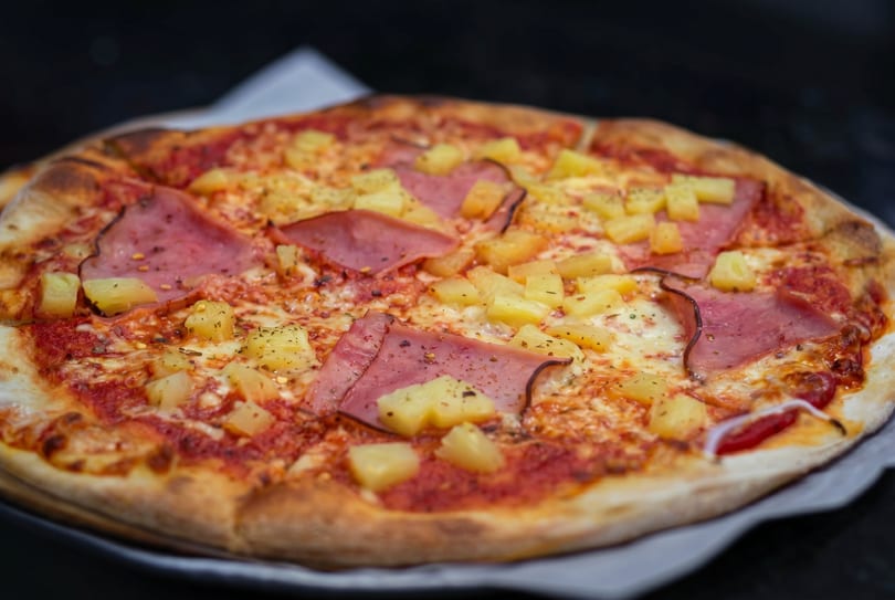 A Hawaiian pizza with ham, pineapple, and melted cheese on a dark background.