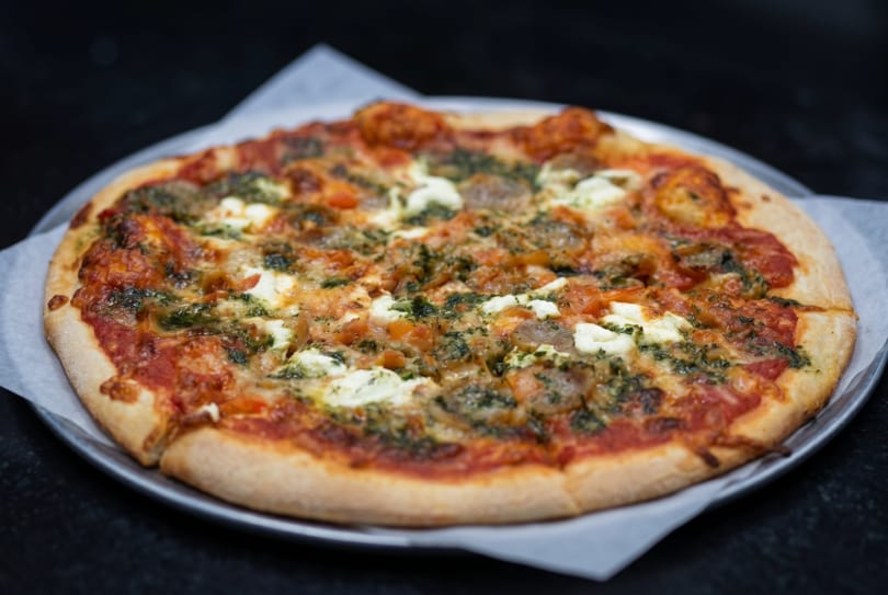 A whole pizza with red sauce, pesto, sausage, and ricotta cheese on a metal pan.